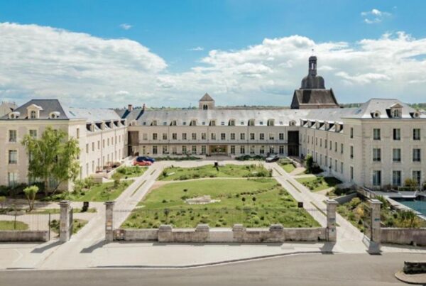 This is an image of a large castle complex with a massive building and a green field in the centre full of people.