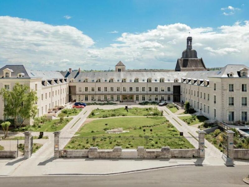 This is an image of a large castle complex with a massive building and a green field in the centre full of people.