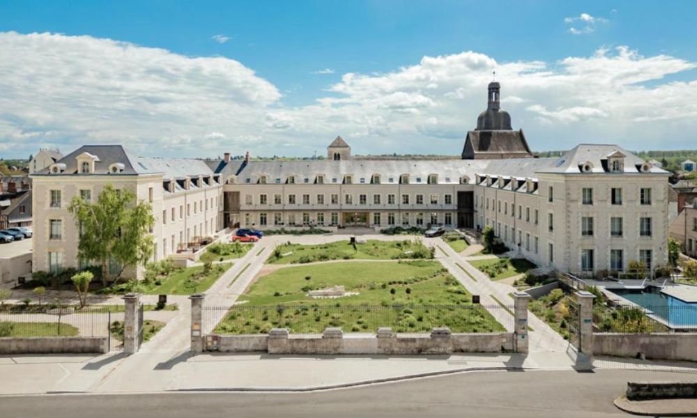 This is an image of a large castle complex with a massive building and a green field in the centre full of people.