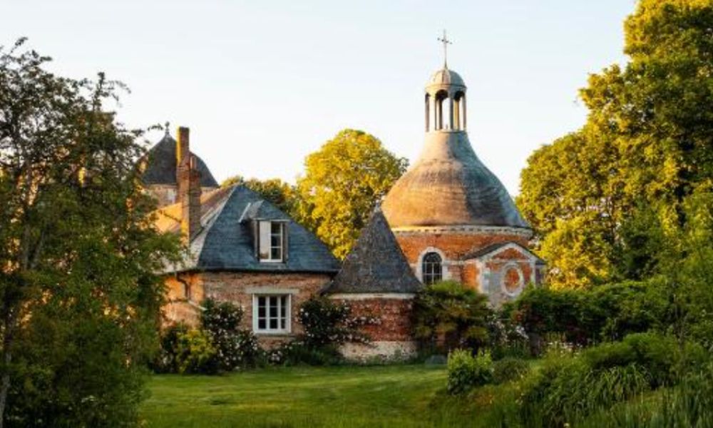 This is an image of a small red brick cottage house behind a clearing of trees.
