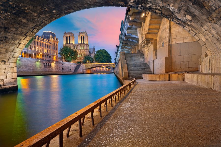 This is an image of a bridge with water underneath from the canal and Seine, with a sunset peeping through.