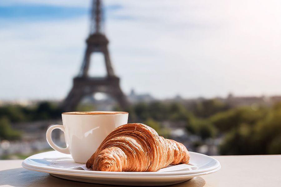 This is an image of a croissant and a coffee with a view of the Eiffel Tower behind it.