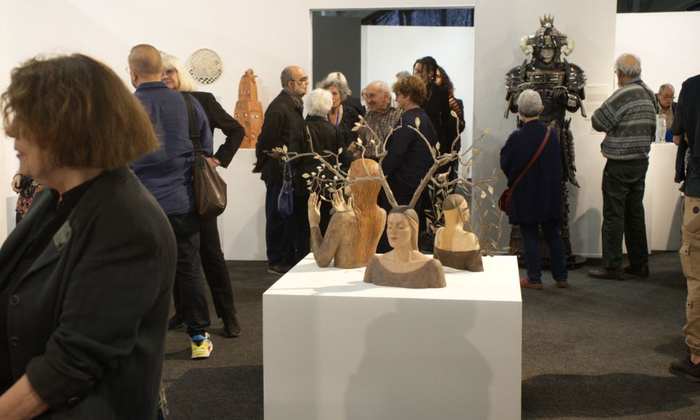 This is an image of some model heads on a plinth in the middle of an art exhibit that people are walking around.