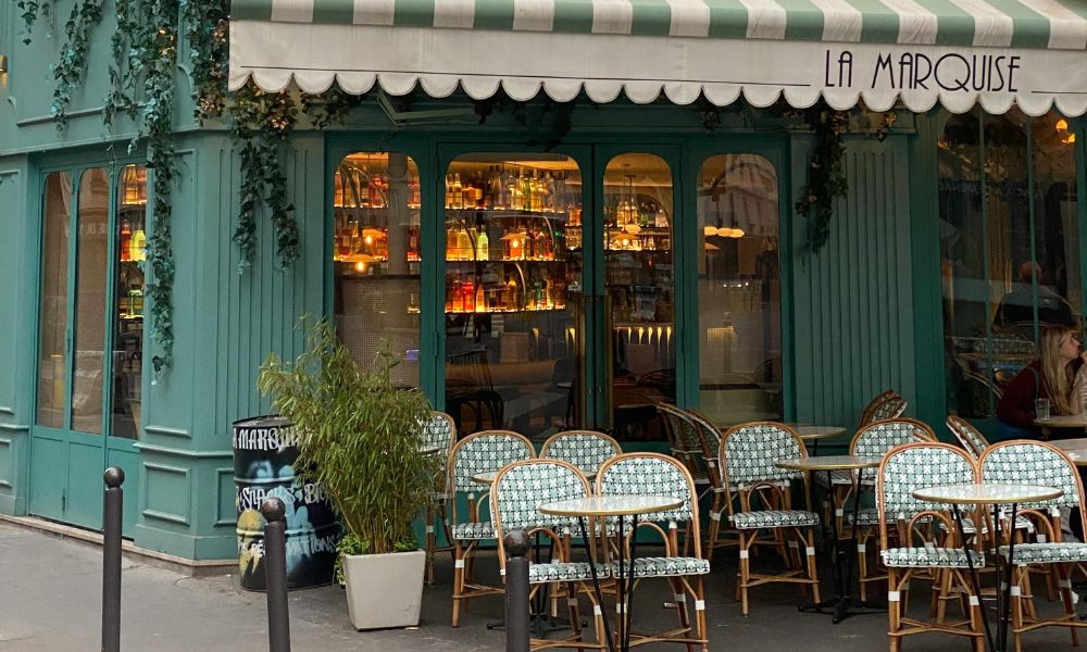 This is an image of a Parisian cafe with tables and chairs outside and a nice seating area inside its glass doors.