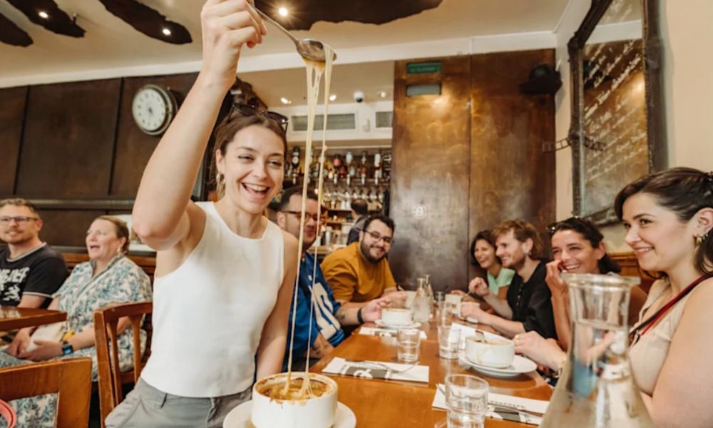 This is an image of a group of people on a food tour eating food and there is one girl doing a massive cheese pull from the French onion soup.