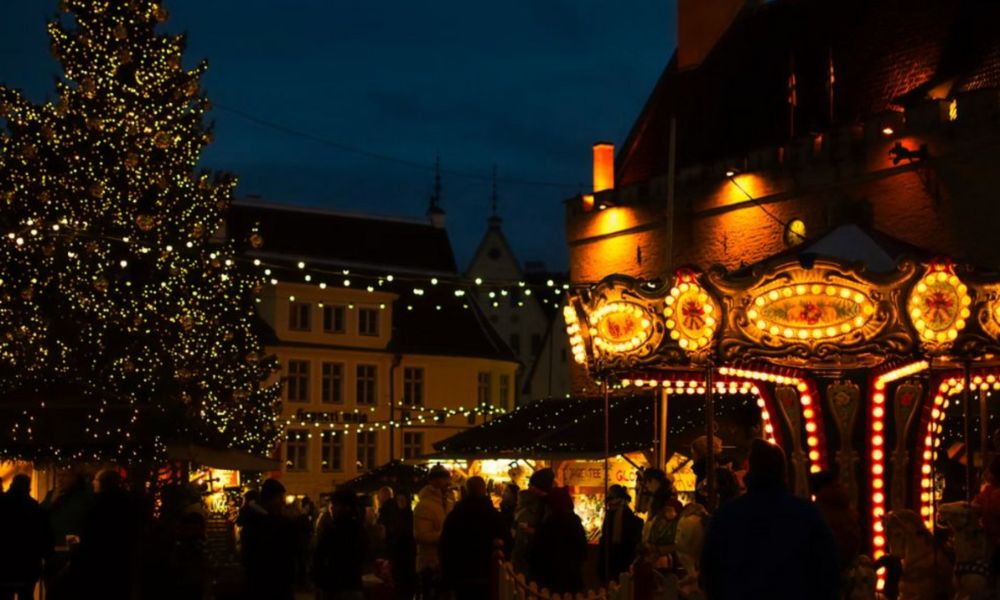 This is an image of a little Christmas village lit-up with a merry-go-round, big christmas tree and lots of people milling about.