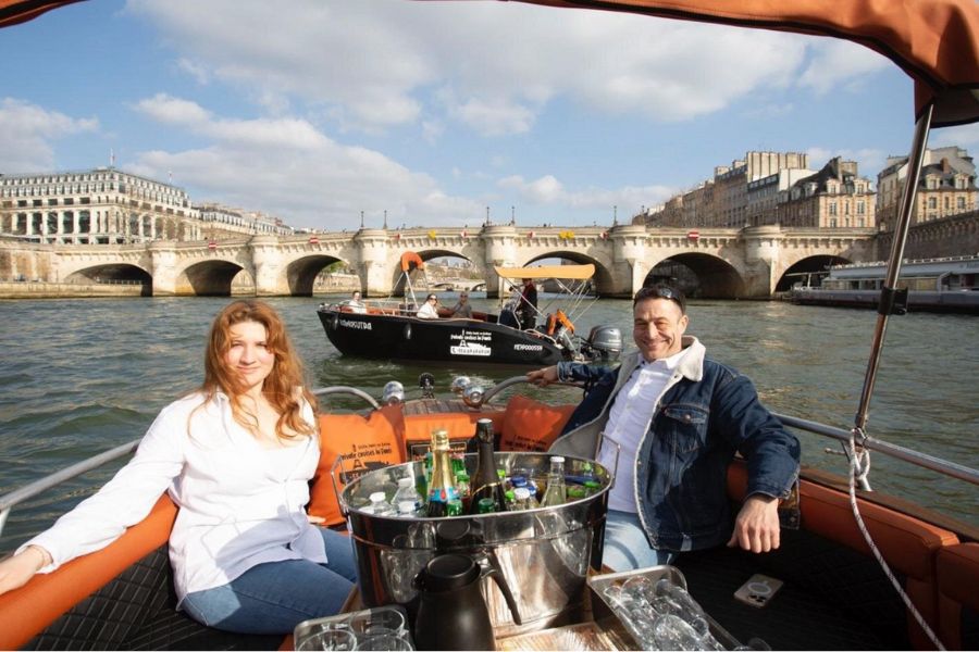 This is an image of a woman and man sitting in a private tour boat with drinks on the Seine with a bridge behind them.