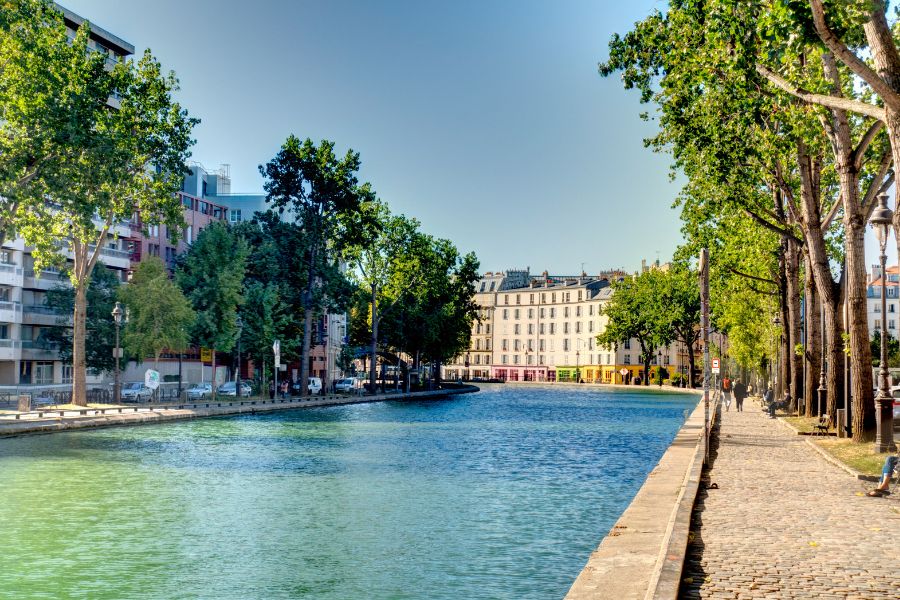A calm, tree-lined stretch of Canal Saint-Martin on a clear summer's day, with cobblestone towpaths, Haussmann-style buildings, and colourful shopfronts reflected in the still green water.