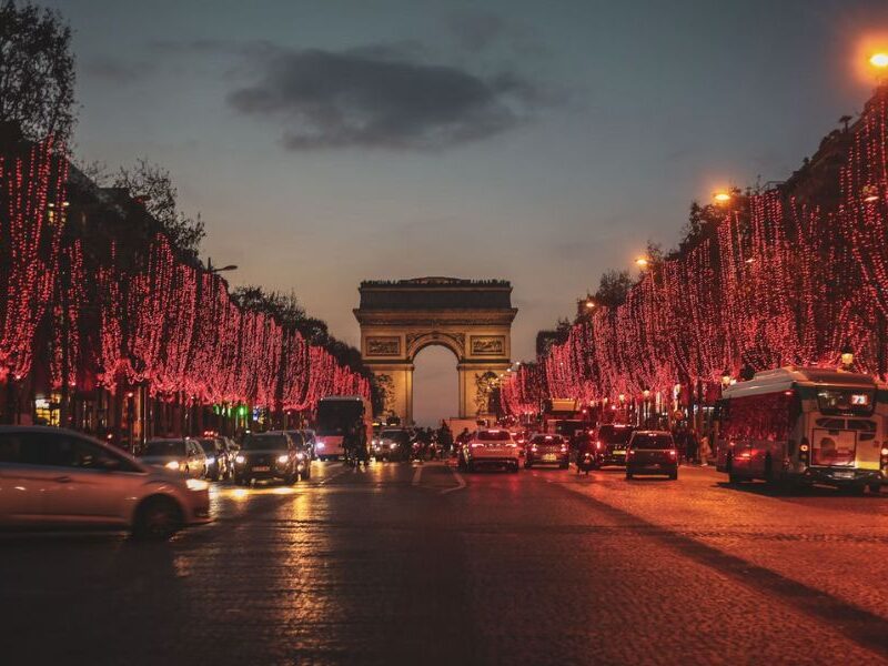 This is an image of the Arc de Triomphe with trees lining the street leading up to it, which are all dressed with red Christmas lights.