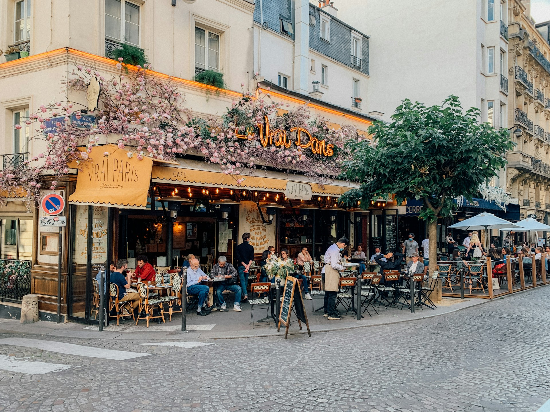 A busy cafe on the corner of a Parisian street. Flowers adorn the walls outside.