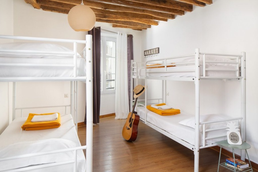 This is an image of a bright hostel bedroom with multiple white bunk beds and orange blankets neatly folded on top. The floor is wooden and there is a guitar by the side of one of the beds.