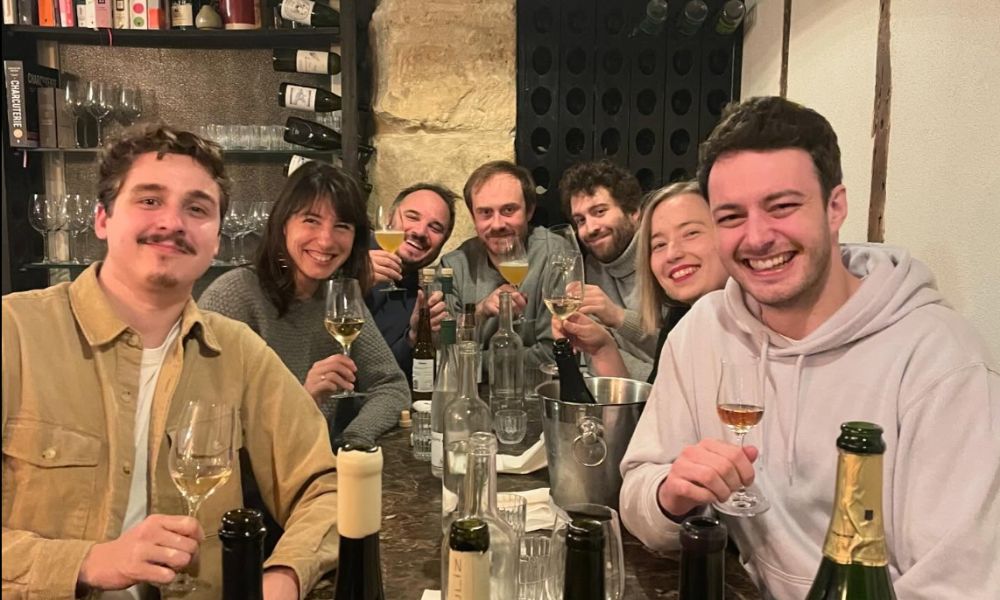 This is an image of a group of people smiling and laughing on a table with wine bottles and posing for the camera.