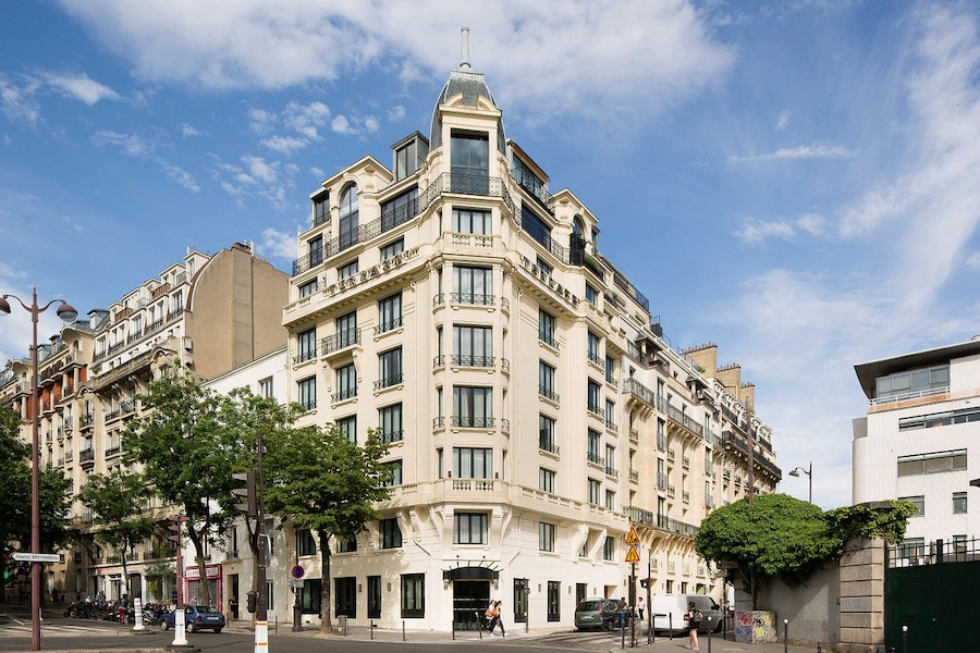 This is an image of the front of a luxurious looking hotel in Paris with clean walls. It is a tall building with lots of windows on it and the sky is bright and blue in the background.
