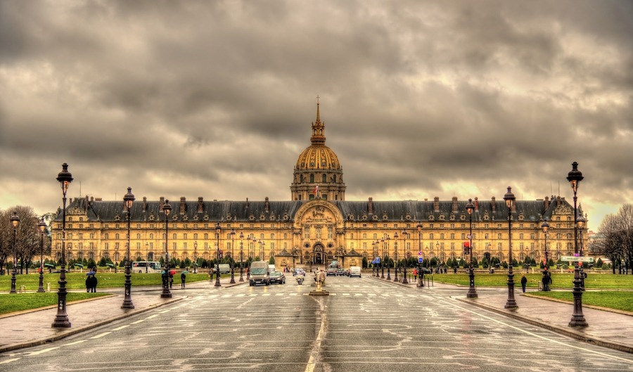 This is an image of Les Invalides in Paris. There are lots of clouds in the background but the front of the building looks well lit and warm in tone. It is extremely grand and big.