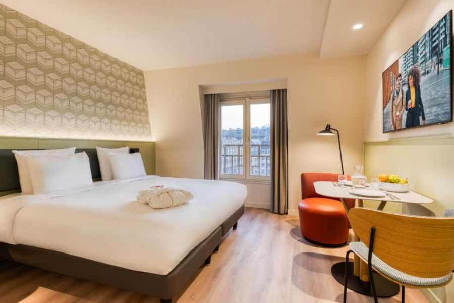 This is an image of a minimalist room with a double bed, desk and couch with lamp. There is also a door that leads out to a balcony that overlooks a view of Montorgueil.