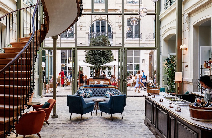 This is an image of the inside of a luxurious hotel looking out of the big glass french windows to the courtyard where people are standing and sitting.