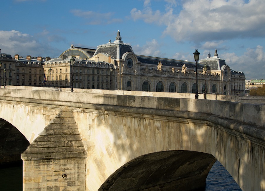 This is an image of the outside of the Musee d'Orsay in Paris. A bridge is also in view in the foreground and a blue sky behind the museum.