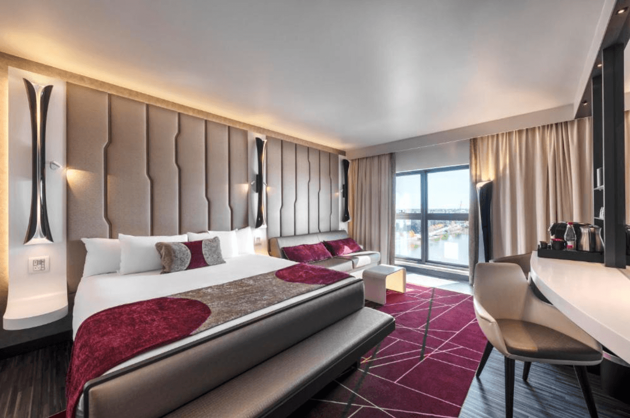 A luxurious hotel room at Disney Hotel New York – Art of Marvel, featuring a large king bed with deep red and taupe bedding, a tall padded headboard, geometric crimson carpet, and floor-to-ceiling windows overlooking a waterfront view.