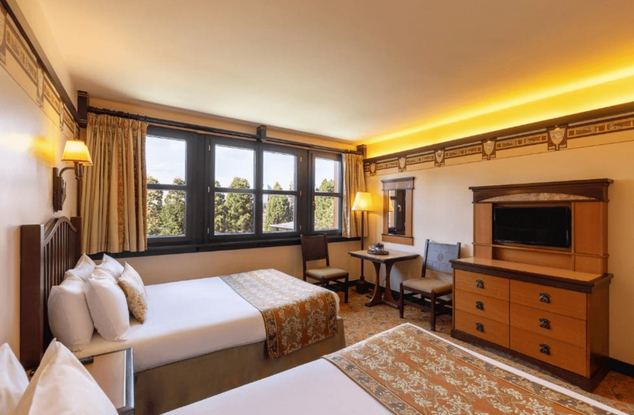 A twin-bedded room at Disney Sequoia Lodge with warm amber lighting, rustic wooden furniture, floral-patterned bed runners, decorative border wallpaper, and large windows overlooking trees.