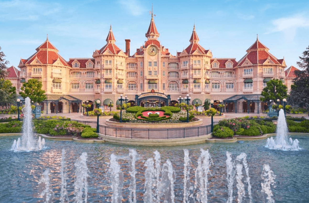 The Disneyland Hotel at Disneyland Paris, a grand Victorian-style pink building with red turrets and a central clock tower, viewed from across an ornamental fountain pool with topiary Mickey Mouse ears in the foreground garden.