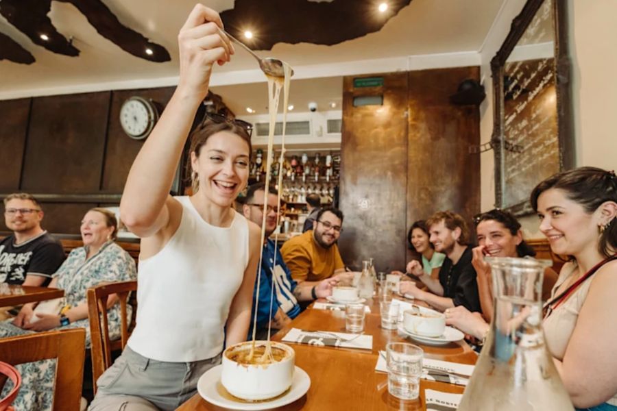 This is an image of a tour group sitting at a table trying french onion soup. The tour guide is using a fork to show everyone else the cheese pull.