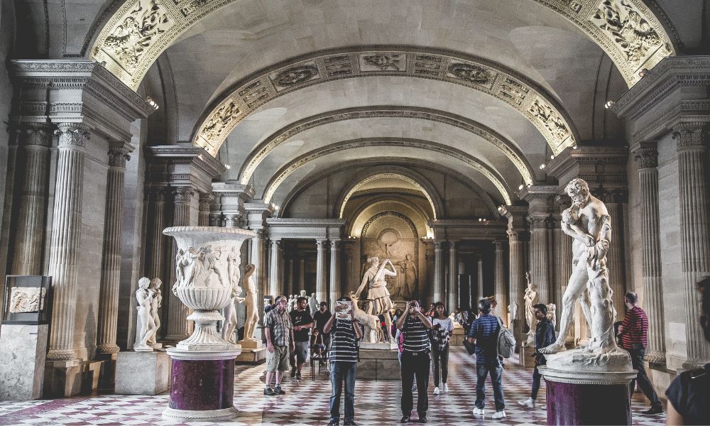 This is an image of the interior of the Louvre with people moving around the room staring up at the many statues.