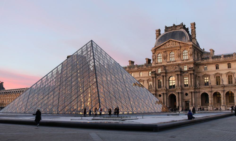 This is an image of the Louvre with a pink sunset behind it and a golden building.