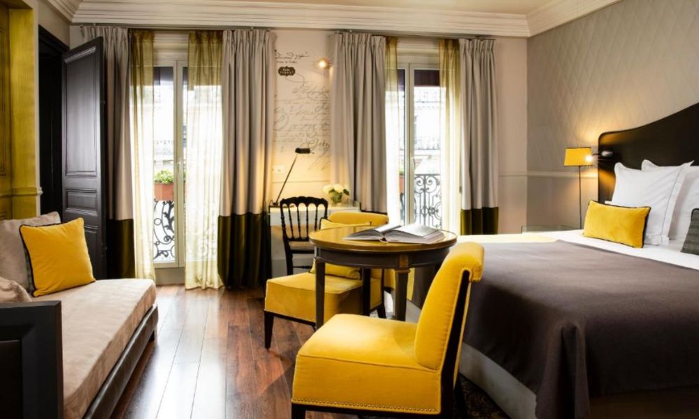 This is an image of a big, spacious hotel room with a big bed and a small table at the end of it. There is also a couch along the wall. The colours are brown with accent yellows.