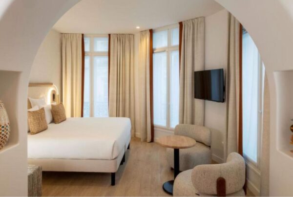 This is an image of a big, spacious hotel room with a white bed a small table under a tv on the wall. There are floor to ceiling windows with curtains.