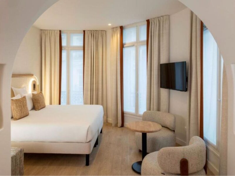 This is an image of a big, spacious hotel room with a white bed a small table under a tv on the wall. There are floor to ceiling windows with curtains.