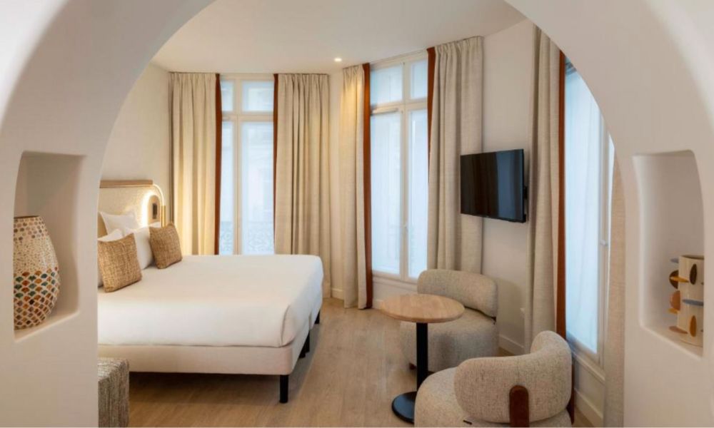 This is an image of a big, spacious hotel room with a white bed a small table under a tv on the wall. There are floor to ceiling windows with curtains.