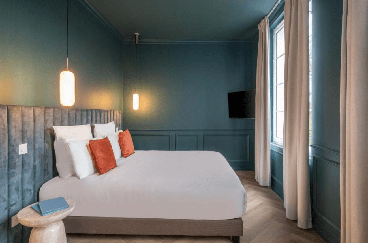 A boutique hotel room at L'Horloge Aparthotel with deep teal walls and ceiling, a velvet upholstered headboard, white bedding with terracotta accent cushions, herringbone wood flooring, and pendant lights.
