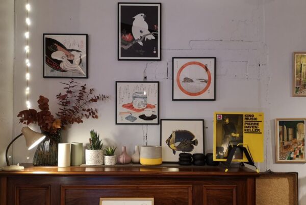 This is an image of a bunch of printed artworks framed and put on the wall above a chest of drawers with candles and plants on top of it.