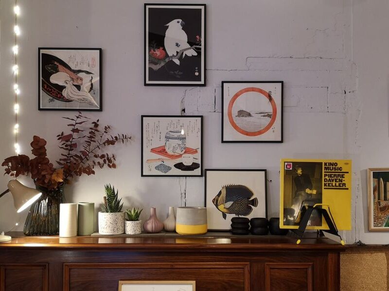 This is an image of a bunch of printed artworks framed and put on the wall above a chest of drawers with candles and plants on top of it.