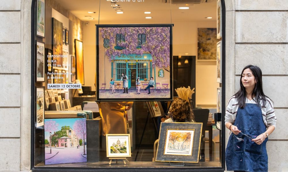 This is an image of a lady standing out front of a print shop, with a big window full of framed artwork.