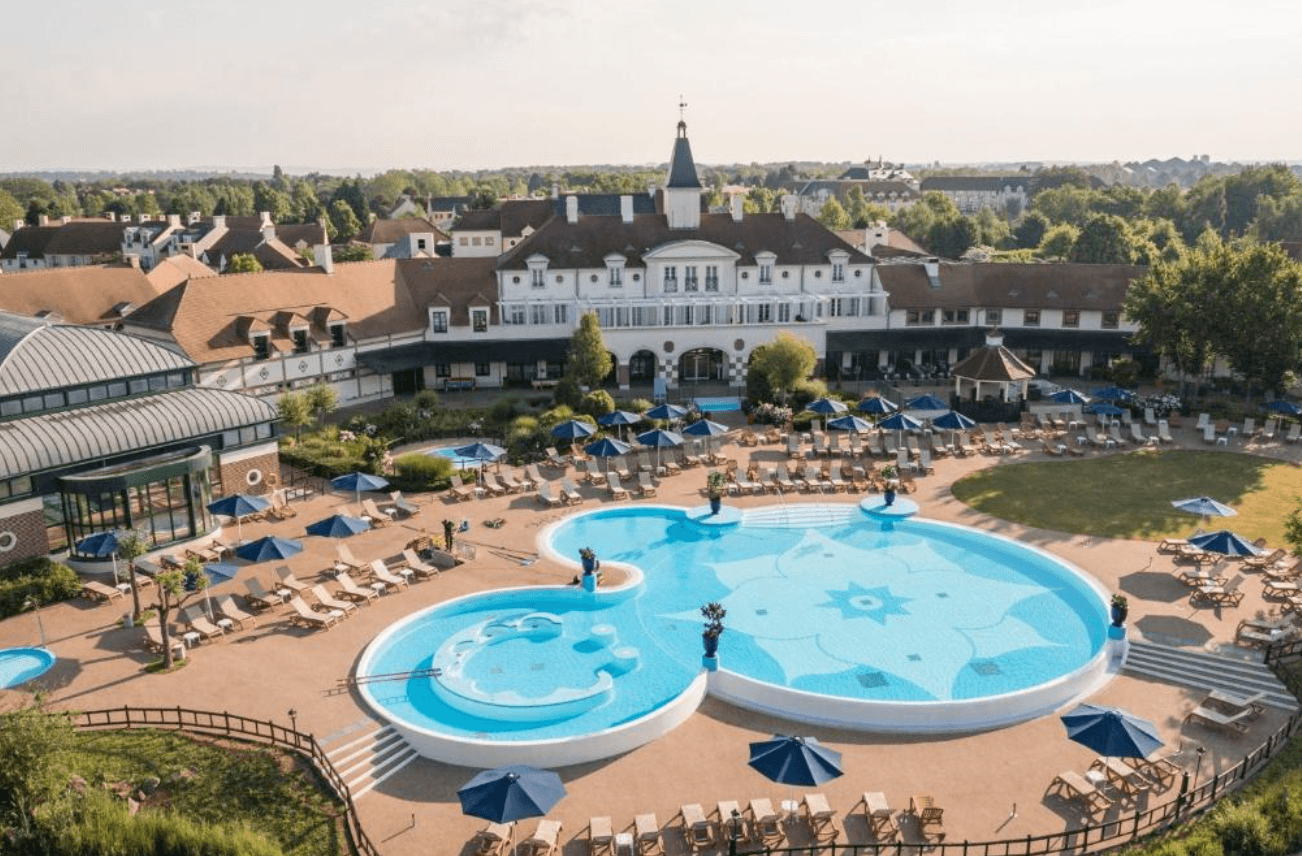 Aerial view of Marriott's Village d'Île-de-France showing a Mickey Mouse-shaped outdoor swimming pool surrounded by sun loungers and blue parasols, with the white château-style main building and lush grounds behind.