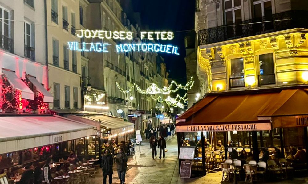 This is an image of a decorated street in Paris with plenty of lights up and cafes + bars that have tables and chairs spilling out into the street.