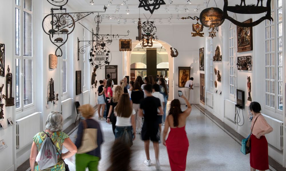 This is an image of a big hall room with people walking through it looking at beautiful works on the walls. There are cool metal lighting pieces and fixtures coming down from the ceiling.