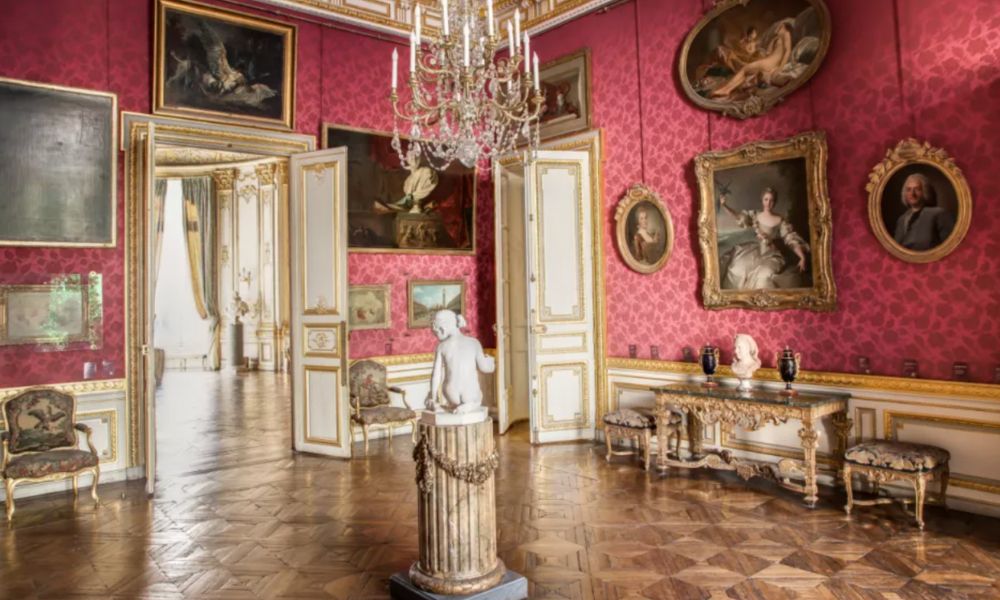 This is an image of a big room with a bunch of incredible renaissance art hung around and a massive chandelier in the centre that is directly over a statue.