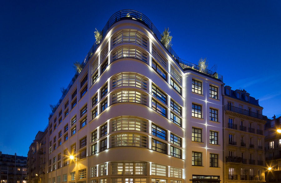 This is an image of a beautiful sleek and modern hotel front. The sky is a deep dark blue in the background and it looks like mid evening time. The hotel is also lit in a sleek way.