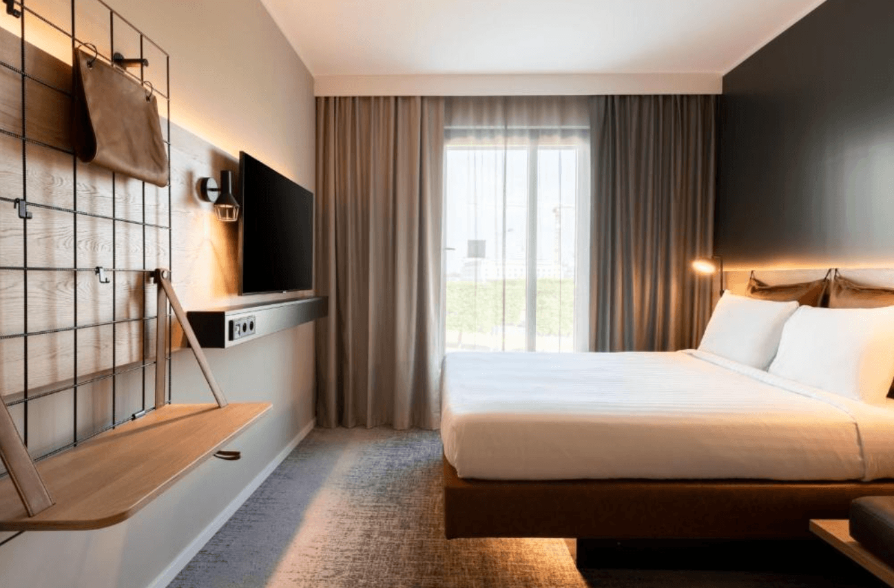 A modern hotel room at Moxy Paris Val d'Europe with a warm wood and black industrial aesthetic, wall-mounted TV on a timber panel, a fold-down desk with metal grid shelving, and a queen bed with crisp white bedding.