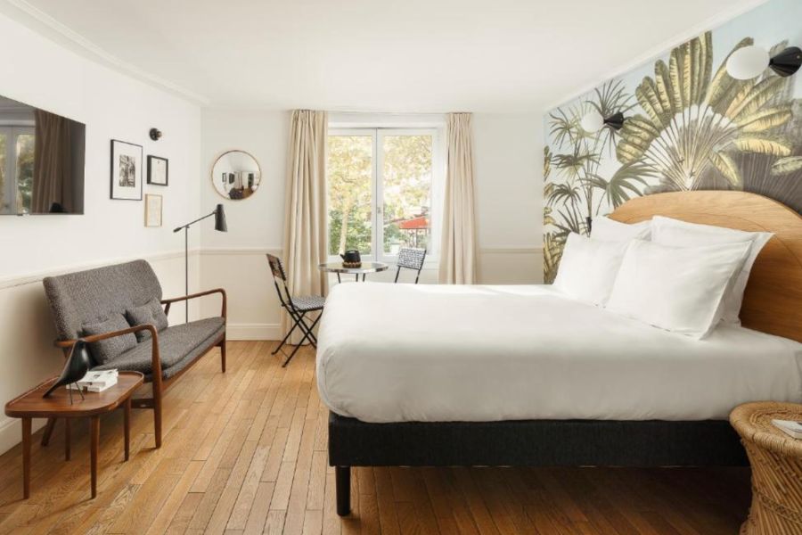 This is an image of a minimalist hotel room wiith a bright open window, a double bed and a small couch in front of the bed. There is also a big feature wall with a picture of a plant.