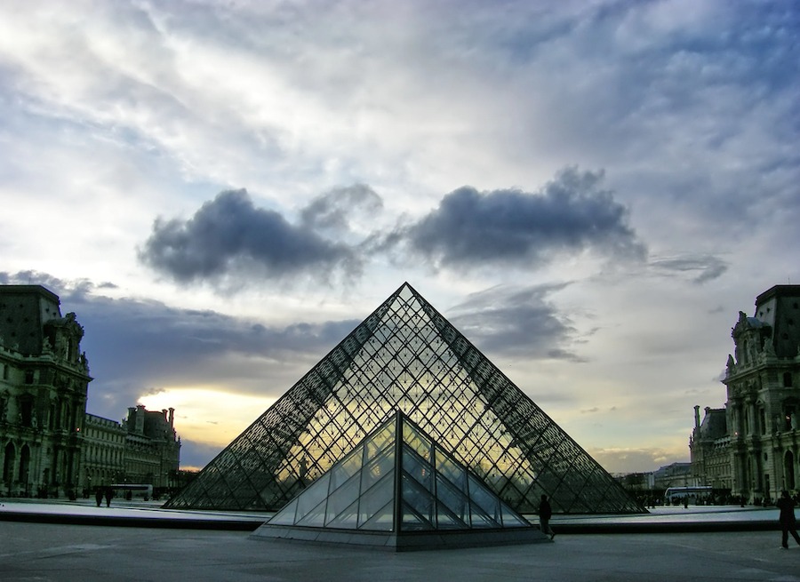 This is an image of the glass triangle entrance to The Louvre museum in Paris. The sky is full of pretty clouds in the background and it is dusk.