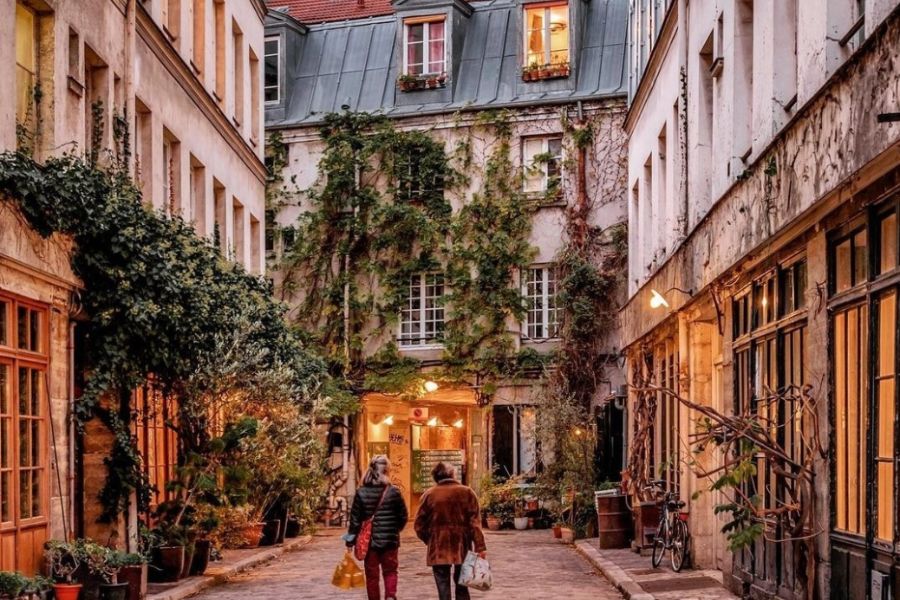 This is an image of a green street with lit-up shops lining the way down. There are two people leisurely walking home in the centre.