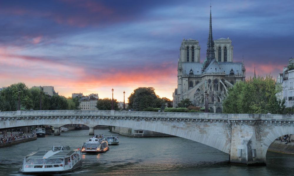 This is an image of a bridge in Paris with boats going underneath it across the Seine. Behind it, you can see the Notre Dame and a beautiful pink sunset.