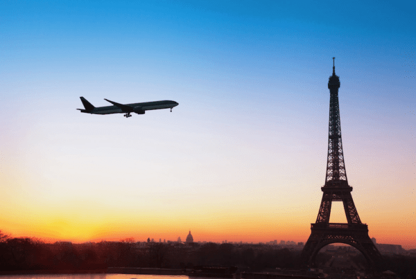 This is an image of a plane flying close to the Eiffel Tower. In the background the sun is setting and the sky has lovely soft colours of blue, orange and yellow. It is dreamy and picturesque.