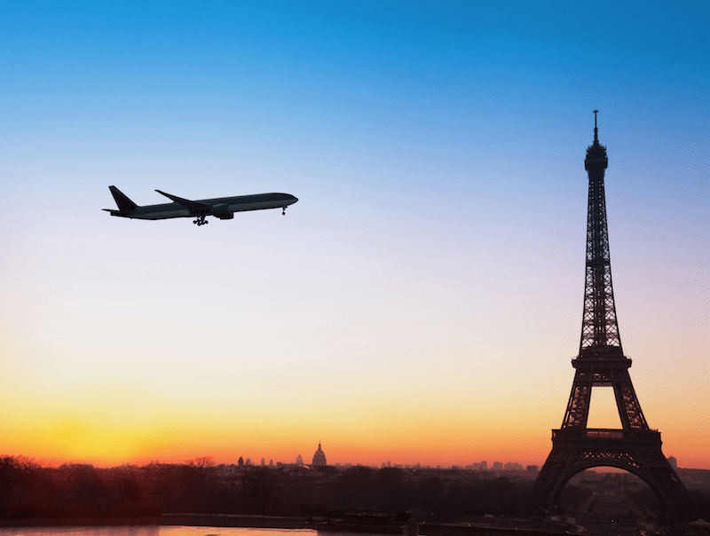 This is an image of a plane flying close to the Eiffel Tower. In the background the sun is setting and the sky has lovely soft colours of blue, orange and yellow. It is dreamy and picturesque.