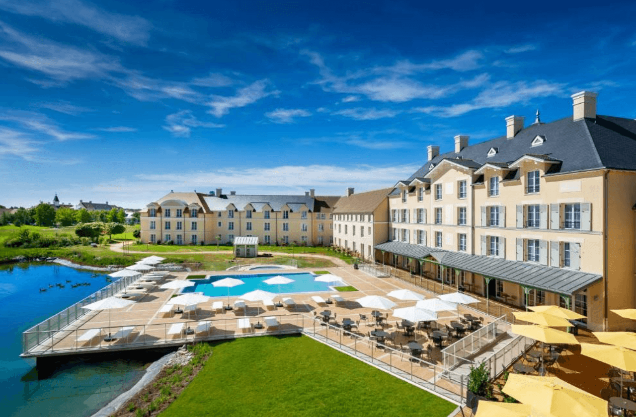 Aerial view of Staycity Aparthotels near Disneyland Paris, a cream-coloured French manor-style property with a rectangular outdoor pool, white parasols, lakeside terrace, and manicured gardens under a bright blue sky.