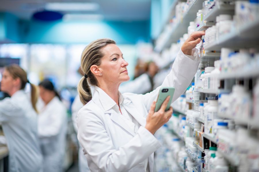 A female pharmacist in a white lab coat reaches up to a shelf stocked with products while holding a smartphone, with other staff members visible working in the background of a well-stocked pharmacy.