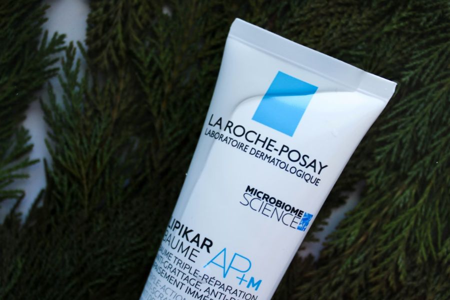 A tube of La Roche-Posay Lipikar Baume AP+M moisturising balm, labelled "Microbiome Science," resting against a backdrop of green pine branches.